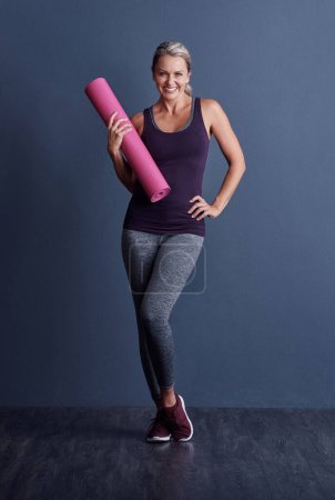 Foto de El yoga es más beneficioso de lo que la mayoría piensa. Retrato de estudio de una atractiva mujer madura en ropa deportiva sosteniendo una alfombra de ejercicio sobre un fondo azul - Imagen libre de derechos