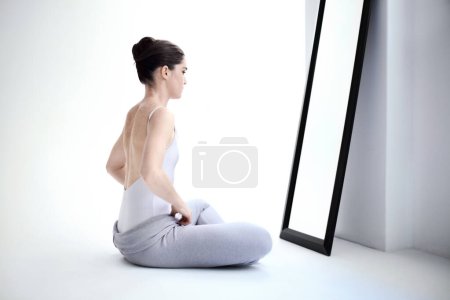Photo for Female ballerina stretching by a mirror in a studio before a dance class, performance or concert. Art, creative and back of young ballet dancer doing a body warm up for classical dancing in her room - Royalty Free Image