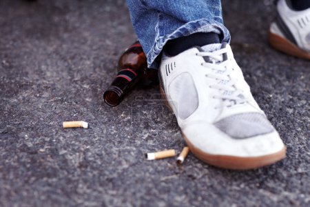 Foto de Pies, cigarrillos y cerveza en un camino de la ciudad para un hombre con zapatillas que fuma y bebe. Zapatos, adicción y primer plano del tabaco y una botella de alcohol en la calle al aire libre de una ciudad urbana - Imagen libre de derechos