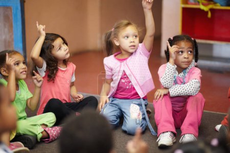 Téléchargez les photos : Éducation, maternelle et enfants posant une question les mains levées alors qu'ils étaient assis sur le sol d'une salle de classe pour le développement de l'enfant. École, apprentissage ou curiosité avec les enfants en classe pour étudier la croissance. - en image libre de droit