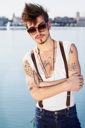Photo for Sunglasses, portrait and man by sea with arms crossed for stylish tattoo, body art and fashion. Hipster, ocean and confident male person standing outdoor in Spain with cool clothes, attitude and punk. - Royalty Free Image