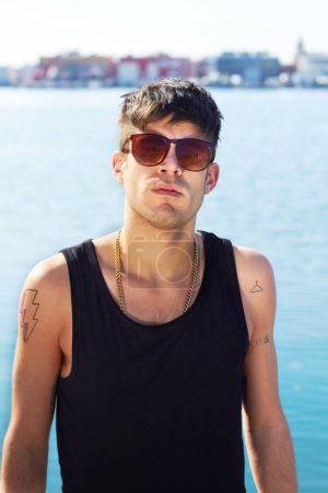 Photo for Portrait, sunglasses and serious man by ocean with stylish tattoo, body art and fashion. Face, sea and trendy male person standing outdoor in Norway with cool clothes, attitude and youth aesthetic - Royalty Free Image