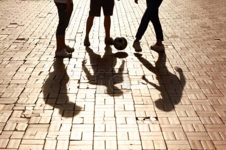 Photo for Sports, football and shadow of friends with a ball training for a skill, trick or stunt on a pavement. Silhouette, soccer and legs of people practicing for a game, match or tournament in the city - Royalty Free Image