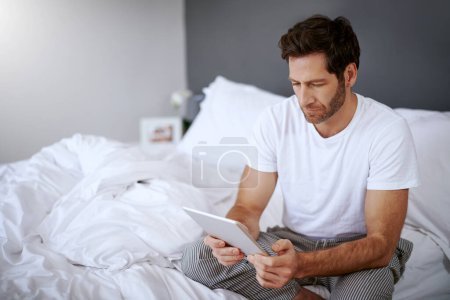 Foto de Buscando remedios para el insomnio. un hombre guapo de mediana edad usando su tableta digital mientras se relaja en su dormitorio - Imagen libre de derechos