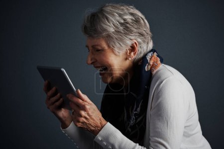 Téléchargez les photos : C'est encore mieux qu'un portable. Plan studio d'une femme âgée joyeuse debout et regardant droit dans sa tablette avec un sourire - en image libre de droit