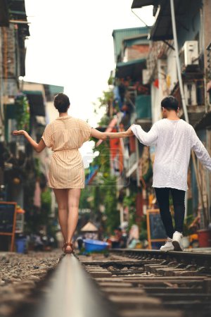 Foto de El amor te hace sentir como un niño de nuevo. Vista trasera de una joven pareja caminando en las vías del tren a través de las calles de Vietnam - Imagen libre de derechos