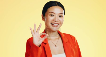 Téléchargez les photos : Sourire, ok signe et le visage de la femme asiatique sur fond jaune pour parfait, bon travail et d'accord. Modèle Emoji, geste de la main et portrait d'une fille heureuse en studio avec succès, symbole d'accord et icône oui. - en image libre de droit