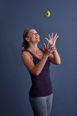 Foto de Ser saludable no tiene que ser una lucha. Estudio de una atractiva mujer madura lanzando una manzana sobre un fondo azul - Imagen libre de derechos