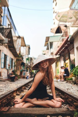 Foto de Viajar debería rejuvenecerte y castigarte. una mujer que lleva un sombrero cónico mientras está sentada en una vía férrea - Imagen libre de derechos