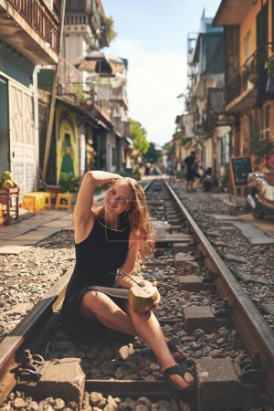 Foto de Disfrutando de los refrescos dulces más populares de las ciudades. una mujer tomando agua de coco mientras se relaja en las vías del tren en las calles de Vietnam - Imagen libre de derechos