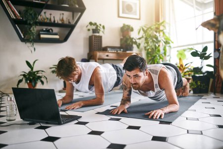 Foto de La fuerza proviene de una base sólida. dos hombres usando un ordenador portátil mientras pasan por una rutina de yoga en casa - Imagen libre de derechos