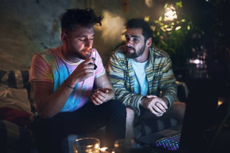 Foto de Es hora de perseguir lo alto. un joven guapo encendiendo un porro de cannabis mientras está sentado con su amigo en casa - Imagen libre de derechos