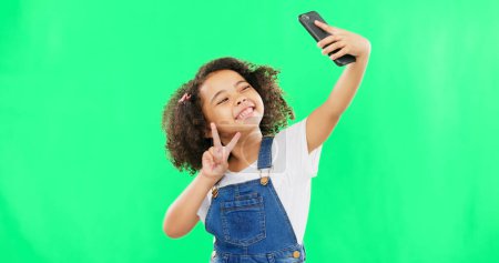 Phone, selfie and child peace sign in a studio with happiness from profile picture. Isolated, green background and young girl with technology showing a v hand gesture for social media with a smile.