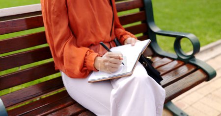 Foto de Parque, banco y una mujer escribiendo en un cuaderno, diario o diario mientras está al aire libre en un jardín para relajarse. Manos, inspiración y periodismo con una escritora sentada fuera en un entorno natural. - Imagen libre de derechos