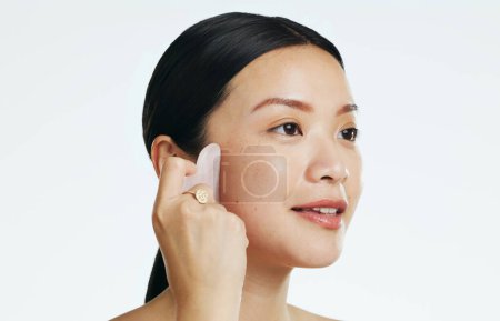Photo for Gua sha, beauty and woman massaging face isolated on a white background in a studio. Spa, skincare and an Asian girl with a product to massage for facial relaxation, treatment and anti aging. - Royalty Free Image