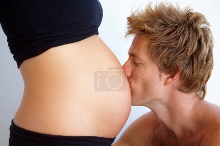 Téléchargez les photos : Homme embrassant l'estomac, la femme enceinte et l'amour, excité de nouveaux parents en studio sur fond blanc. Mariage, soutien et grossesse, mère et père avec bébé dans le ventre et couple avec la future famille - en image libre de droit