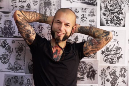 Foto de Tatuajes, creatividad y retrato de un artista masculino parado junto a bocetos o dibujos en su estudio. Feliz, sonrisa y la cara de un hombre punk con un borde, funky y body art o tinta creativa tienda de negocios - Imagen libre de derechos