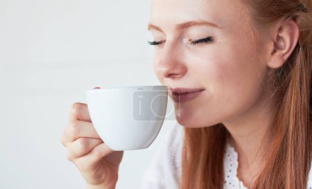 Téléchargez les photos : Visage de femme sentir sa tasse de café dans un studio pour un parfum le matin un week-end. Modèle calme, relaxant et féminin profitant de l'arôme d'un cappuccino, caféine ou latte sur fond blanc - en image libre de droit