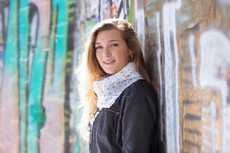 Téléchargez les photos : Portrait, mode et sourire de femme au mur de graffiti en plein air de la ville pour l'art de rue. Visage, style d'hiver et heureux, gen z ou jeune femme de Suède avec écharpe cool, veste en cuir ou vêtements. - en image libre de droit