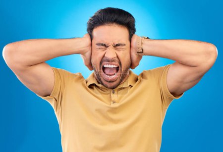 Photo for Man covering his ears while screaming in a studio for angry, upset or mad argument expression. Crazy, shout and young male person with an open mouth for loud voice gesture isolated by blue background. - Royalty Free Image