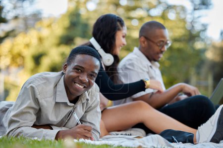Téléchargez les photos : Nature, université et portrait d'un homme étudiant sur l'herbe avec ses amis pour un test ou un examen. Joyeux, sourire et africaine étudiant devoir d'écriture dans son carnet dans le parc sur le campus de l'université - en image libre de droit