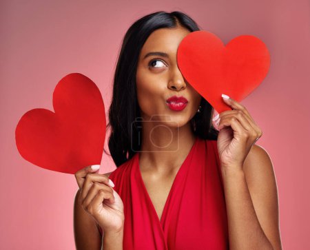 Photo for Red heart, woman in studio and kiss emoji on background for romance, emotion and fun symbol. Happy indian female model, thinking and pout with paper shape for love, icon or flirting on valentines day. - Royalty Free Image