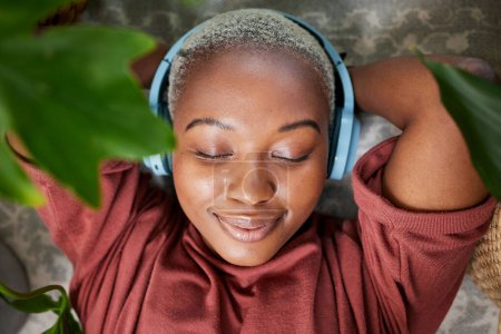 Photo for Woman, face and headphones for listening to music to relax on a floor with plants for peace and calm. Headshot of a black female person meditate with sound, audio and podcast or radio at home. - Royalty Free Image