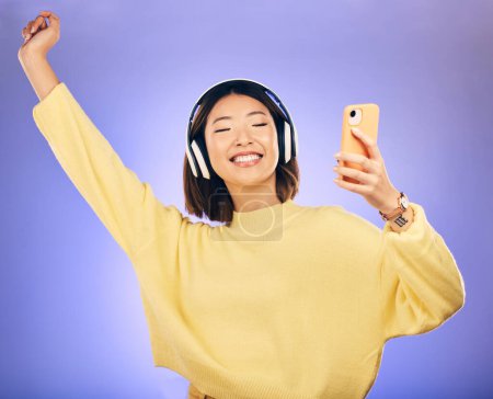 Foto de Danza, música y mujer asiática con teléfono en estudio para streaming, suscripción y radio. Relajarse, auriculares y persona femenina feliz en el teléfono inteligente escuchar audio, canción y pista en el fondo púrpura. - Imagen libre de derechos