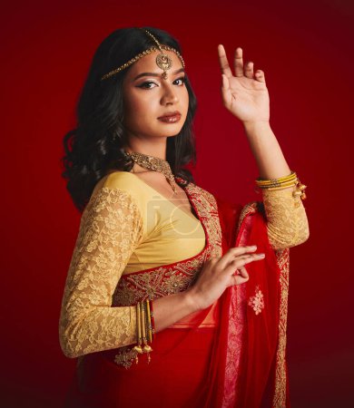 Photo for Portrait, beauty and Indian woman with fashion, traditional dress and luxury against a red studio background. Face, female person and model with cultural clothes, jewellery and gold with religion. - Royalty Free Image