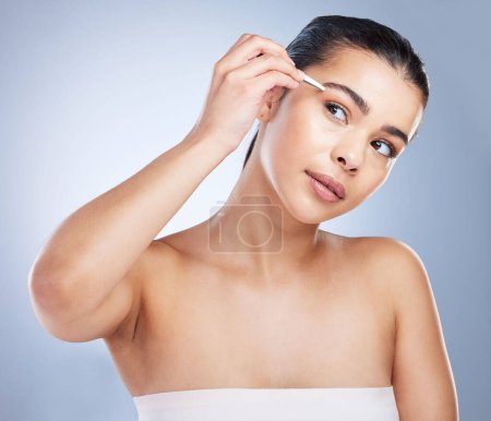 Photo for Young woman tweezing her eyebrows in a studio for grooming or hair removal face routine. Skincare, beauty and female model doing a facial epilation treatment with tweezers isolated by gray background. - Royalty Free Image