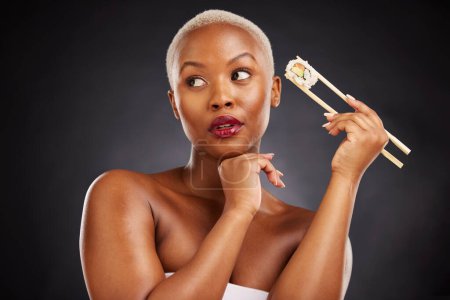 Photo for Face, thinking and woman with sushi and chopsticks in studio for healthy eating, beauty or food. Black female model with makeup idea on dark background for wellness glow, diet or seafood advertising. - Royalty Free Image