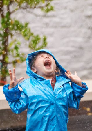 Foto de Clima húmedo, impermeable y una chica jugando en la lluvia al aire libre sola, divirtiéndose durante la estación fría. Niños, invierno o libertad con una adorable niñita con los brazos extendidos afuera. - Imagen libre de derechos