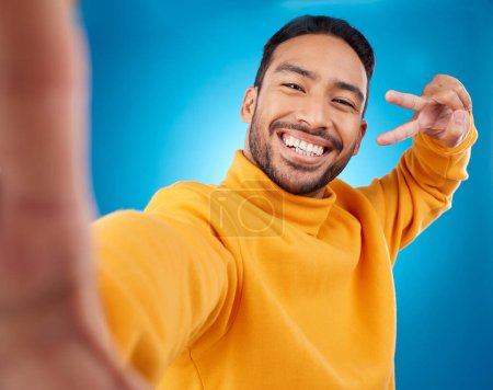 Téléchargez les photos : Selfie, portrait joyeux et paisible d'un homme en studio avec main, emoji et sourire. Homme asiatique modèle de mode sur un fond bleu avec un état d'esprit positif pour les médias sociaux profil photo mise à jour. - en image libre de droit