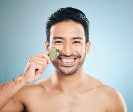 Photo for Face, man and beauty, gua sha and cosmetic tools, smile in portrait and natural treatment on blue background. Skincare, healthy glow and jade stone, dermatology and wellness with male model in studio. - Royalty Free Image