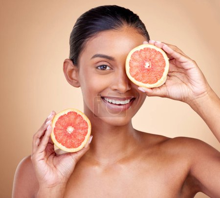 Photo for Beauty, woman and grapefruit on eye for skincare, cosmetics and natural product, health or vitamin c in portrait. Face of indian person or model, red fruit and dermatology on studio, brown background. - Royalty Free Image