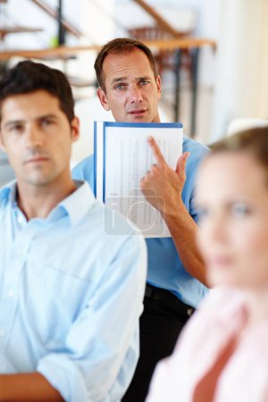 Photo for You mean we have to get up to here. Three colleagues are gathered in an office with one colleague pointing to his notes - Royalty Free Image