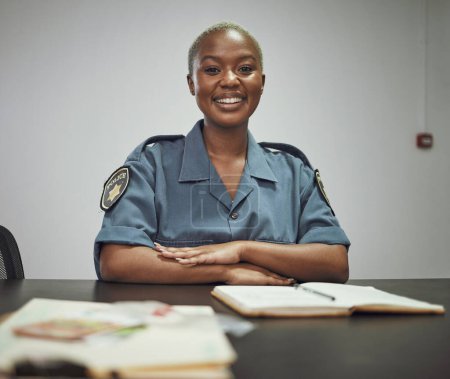 Téléchargez les photos : Joyeux, sourire et portrait d'une policière assise près d'un bureau dans le bureau qui rédige des rapports. Confiance, bonheur et garde de sécurité africaine avec un état d'esprit positif sur le lieu de travail - en image libre de droit