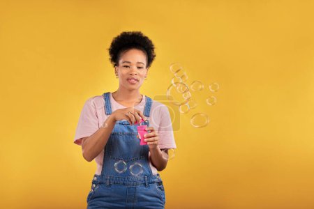 Photo for Woman, blowing bubbles and fun portrait in studio while playful for birthday, space or party celebration. Black female model person with liquid soap, natural beauty and fashion on a yellow background. - Royalty Free Image