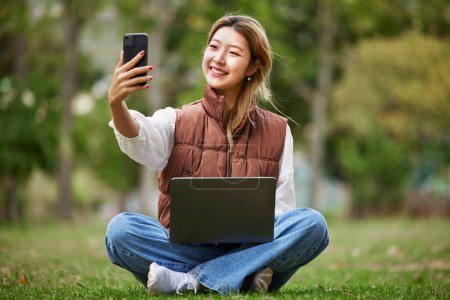 Foto de Selfie, estudiante asiática y mujer en videollamada en un parque para conectarse al contacto mientras estudia al aire libre en línea. Internet, ordenador portátil y joven o influencer toma fotos en la naturaleza para la paz y la calma. - Imagen libre de derechos