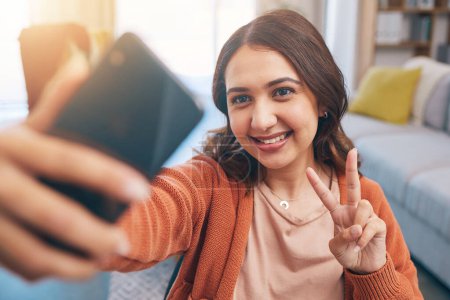 Photo for Face, woman and peace for selfie with smile in home while live streaming on video call in living room. Profile picture, happy female gen z and show v sign, hand emoji and memory for social media post. - Royalty Free Image