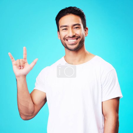 Photo for Man, horns and love in studio portrait with smile, kindness and sign language by blue background. Young guy, student and happy with icon, emoji and hand with care, romance or vote with white t-shirt. - Royalty Free Image