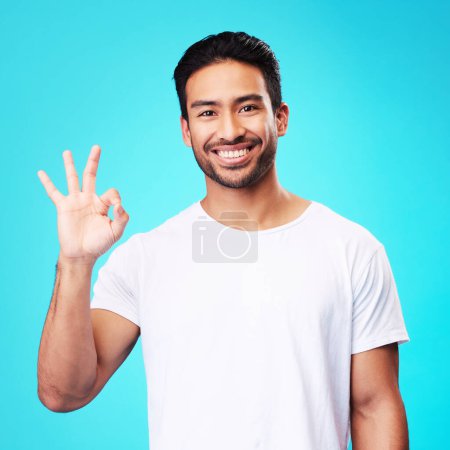 Photo for Happy, perfect gesture and portrait of a man in a studio with an agreement sign or expression. Happiness, smile and young Indian male model with an approval hand emoji isolated by a blue background - Royalty Free Image