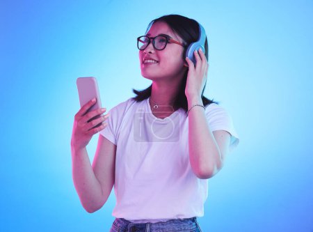 Photo for Woman, student and phone with headphones listening to music, techno streaming and radio on blue background. Person, gen z or youth audio, electronics and mobile subscription in neon light and studio. - Royalty Free Image
