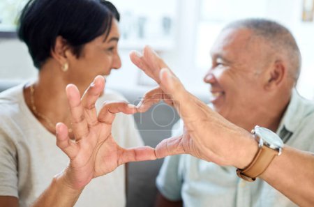 Foto de Forma de corazón, manos y pareja de ancianos uniéndose y hablando en la sala de estar de su casa. Feliz, sonrisa y primer plano de un anciano jubilado con un signo de amor o gesto juntos - Imagen libre de derechos