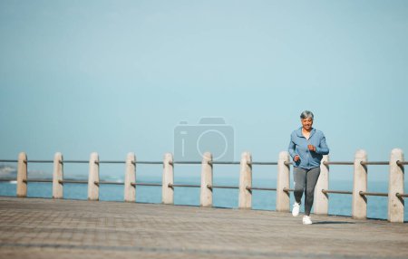 Photo for Fitness, running and senior woman at the beach for exercise, health or cardio on blue sky background. Training, wellness and elderly female runner at the sea for morning workout, routine or ocean run. - Royalty Free Image