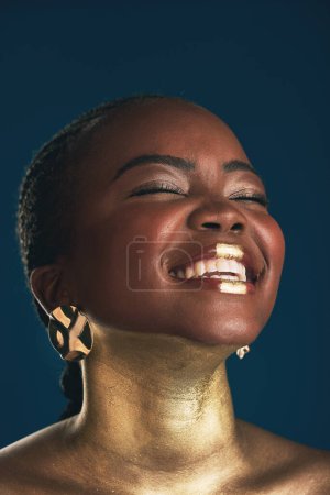 Téléchargez les photos : Maquillage doré, visage et femme noire riant de créativité drôle, peinture cosmétique faciale et design d'art de beauté. Culture africaine blague, comédie ou glamour personne avec visage créatif lueur sur fond bleu. - en image libre de droit