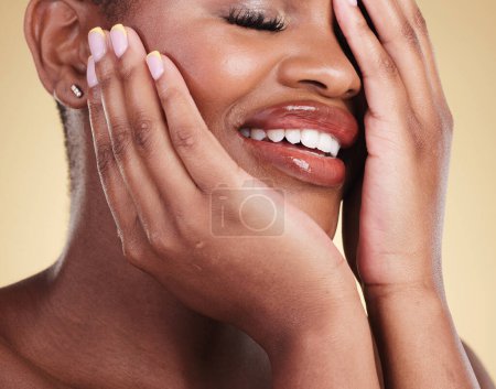 Téléchargez les photos : Mains sur le visage, le maquillage et la beauté de la femme noire en studio pour les soins personnels, l'éclat de la peau ou les cosmétiques. Gros plan de la personne modèle esthétique africaine avec brillance faciale, dermatologie et sourire sur fond beige. - en image libre de droit
