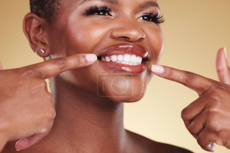 Téléchargez les photos : Sourire, beauté et une femme pointant les dents en studio avec soin de la peau, éclat et maquillage. Visage d'une personne modèle africaine avec brillance dentaire, dermatologie et cosmétique annonce sur un fond beige. - en image libre de droit