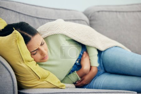 Photo for Stomach pain, pms and woman on a sofa with stress, bad or gut health, gas or ibs in her home. Abdomen, anxiety and female person in living room with gas, digestion or constipation from endometriosis. - Royalty Free Image