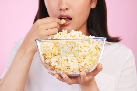 Téléchargez les photos : Popcorn, mains affamées et femmes mangeant une collation de film dans un studio avec regarder la télévision et la nourriture. Fond rose, la personne féminine et la bouche avec des puces dans un bol en verre avec abonnement film et série. - en image libre de droit